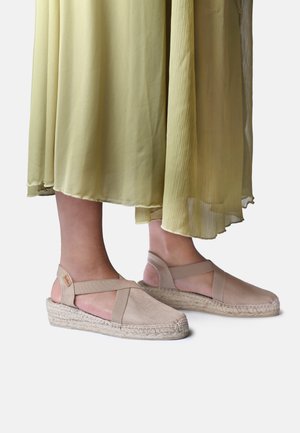 Fødder iført beige espadrille kilehælssandaler med ankelremme, kombineret med en flydende lysegrøn plisseret nederdel.