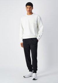 Witte crewneck sweatshirt met een textuurlzak, gecombineerd met zwarte joggingsbroek met twee voorzakken en elastische boorden. Witte sneakers.