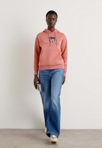 Felpa rosa con cappuccio, tasca frontale e lacci, abbinata a jeans svasati azzurri e sneakers bianche. Modella che tiene un telefono.