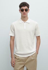 Polo bianca a maniche corte con tessuto strutturato, colletto a costine e patta con un solo bottone, abbinata a pantaloni beige.