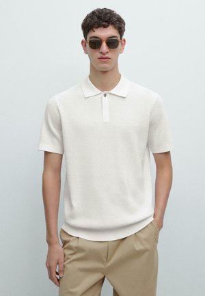 Polo bianca a maniche corte con tessuto strutturato, colletto a costine e patta con un solo bottone, abbinata a pantaloni beige.