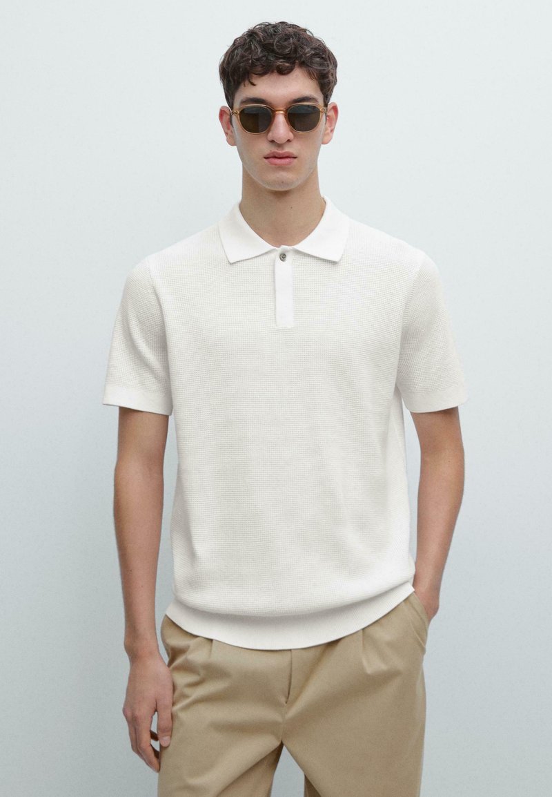 Polo bianca a maniche corte con tessuto strutturato, colletto a costine e patta con un solo bottone, abbinata a pantaloni beige.