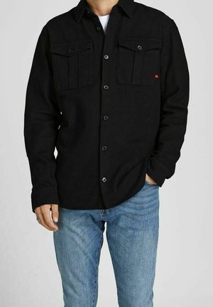 Homme portant une chemise noire boutonnée avec deux poches poitrine boutonnées et un jean bleu clair, une main dans la poche.