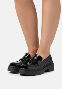 Mocassins en cuir verni noir avec une semelle épaisse, présentant un design élégant, des larges brides et une finition lisse et brillante.