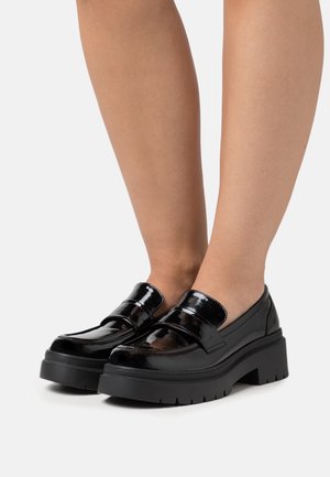 Mocassins en cuir verni noir avec une semelle épaisse, présentant un design élégant, des larges brides et une finition lisse et brillante.