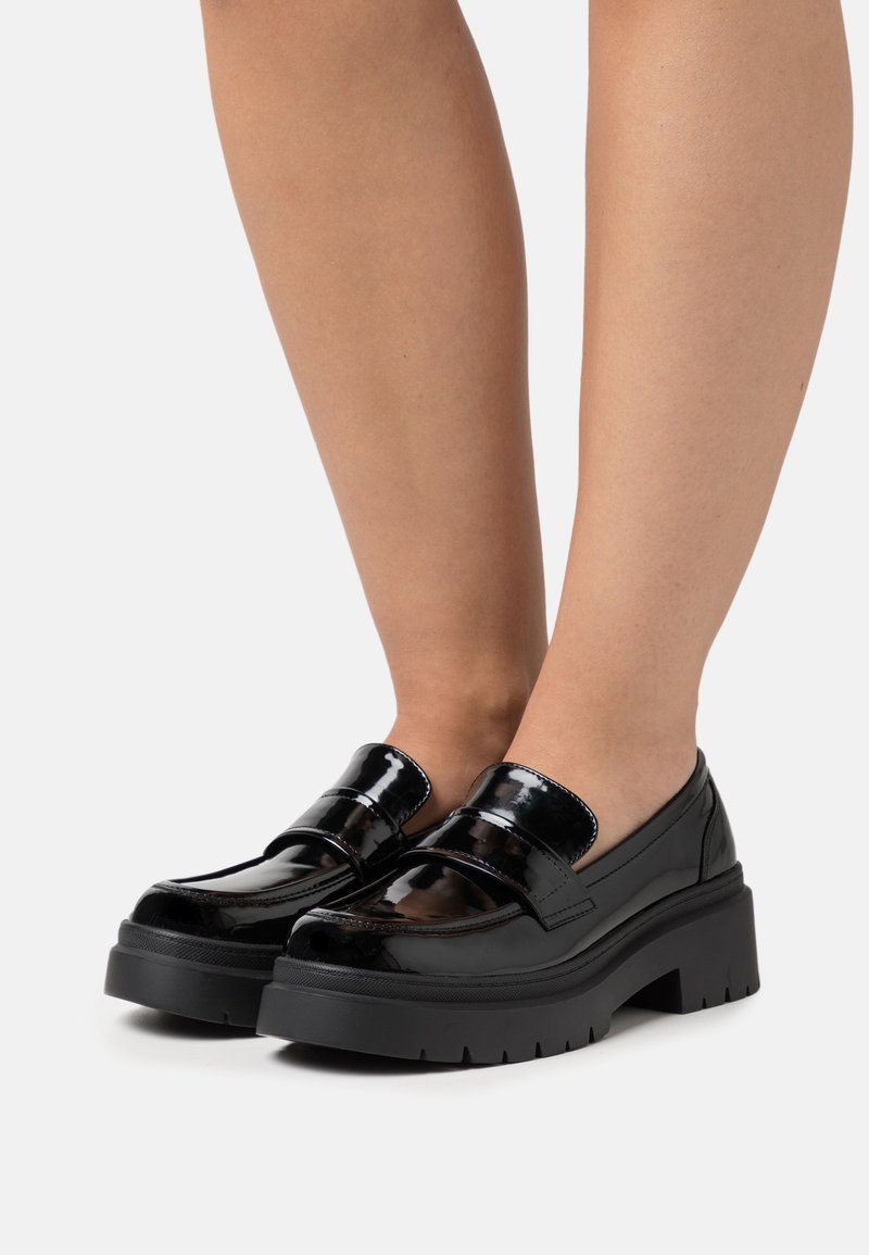 Mocassins en cuir verni noir avec une semelle épaisse, présentant un design élégant, des larges brides et une finition lisse et brillante.