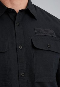 Camisa negra de botones hecha de tela texturizada, que presenta dos bolsillos en el pecho, un parche de logo bordado y botones negros.