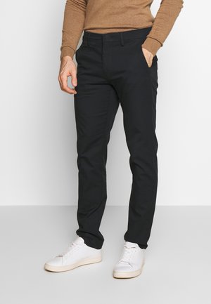 Mann trägt schwarze Hose, weiße Sneakers und ein braunes Langarmshirt, steht mit einer Hand in der Tasche vor einem einfarbigen Hintergrund.