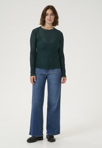 Un haut en tricot à manches longues de couleur vert foncé, avec un design texturé et à motifs ; associé à un jean large bleu et des chaussures noires.