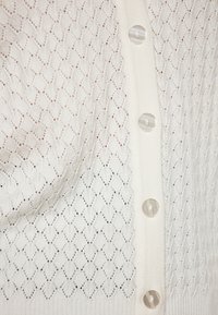 Cardigan alb din tricot, cu un model în diamant, manșete cu nervuri și șase butoane rotunde, translucide, pe toată lungimea închiderii frontale.