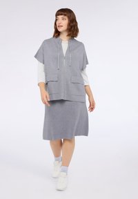 Jasnoszara kamizelka z kapturem i krótkimi rękawami, zapinana na zamek z przodu, z dwoma naszywanymi kieszeniami, zestawiona z dopasowaną spódnicą do kolan. Na nogach białe trampki.