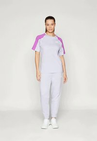 Žena oblečená v svetlo šedom športovom oblečení s fialovými ramennými panelmi a bielymi teniskami, stojaca na svetlom pozadí.