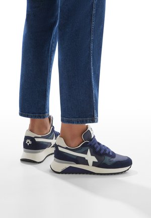 Sneaker blu navy realizzato in materiali scamosciati e in rete, presenta un logo a stella bianca, colletto imbottito e suola testurizzata. Indossato con jeans in denim alla caviglia.