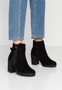 Botines de ante negro con tacón en bloque, paneles laterales elásticos y detalle de hebilla. La suela es texturizada para un mejor agarre.