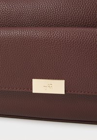 Burgunderfarbene Lederhandtasche mit strukturiertem Finish, ausgestattet mit einer goldenen Metalllogo-Plakette und kontrastierenden Nähten entlang der unteren Kante.