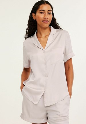 Vrouw met lang krullend haar draagt een lichtgrijs kort mouw blouse met knopen en bijpassende korte broek, staand met handen in de zakken tegen een effen achtergrond.