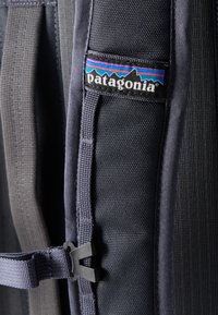 Κοντινή λήψη του λουριού μιας μαύρης τσάντας Patagonia με πολύχρωμο σήμα και ρυθμιζόμενο κούμπωμα.