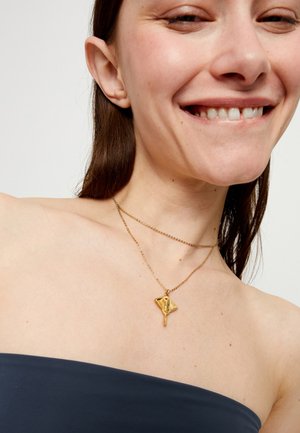 Collar de oro con un colgante en forma de diamante, con un diseño texturizado y cadena colgante, llevado sobre un top azul oscuro sin hombros.