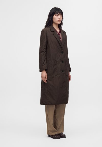 Femme debout portant un long manteau marron foncé, un pantalon large beige et des chaussures foncées sur un fond clair uni.