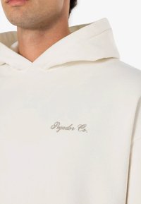 Sudadera color crema hecha de una tela suave, con un bolsillo frontal y un logo bordado en plata que dice "Pegador Co." en el área del lado izquierdo del pecho.