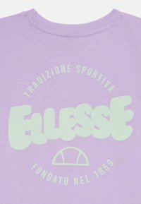 Lavandas T-krekls ar gludu tekstūru, kurā ir liels, piparmētru zaļš logs ar uzrakstu "Ellesse" un teksts izvietots arku formā zem tā.