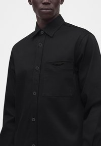 Homme portant une chemise noire à boutons avec une poche poitrine comportant un petit emblème noir sur un fond clair uni.