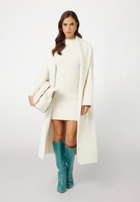 Witte gebreide jurk met een lange witte jas; teal kniehoge laarzen met slangenprint; houdt een beige handtas met zwarte accenten vast.