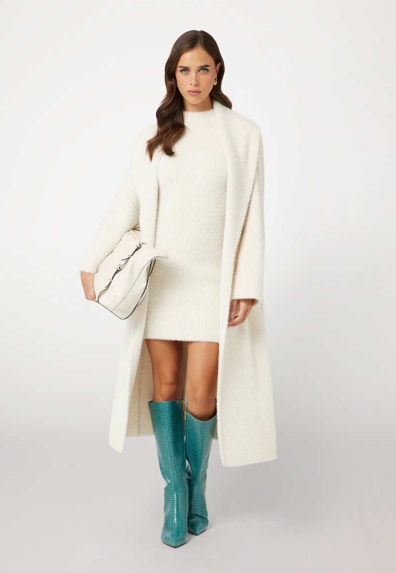 Witte gebreide jurk met een lange witte jas; teal kniehoge laarzen met slangenprint; houdt een beige handtas met zwarte accenten vast.