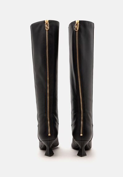 Paire de bottes hautes en cuir noir avec des fermetures éclair dorées courant verticalement dans le dos et des talons bas inclinés sur fond blanc.