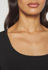 Kvinnas underansikte, hals och övre bröst med medelbrun hudton, iklädd en svart topp med rundad halsringning, mörkbrunt hår synligt på vänster sida.