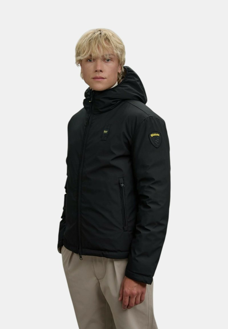 Schwarze isolierte Jacke mit Kapuze, Reißverschluss vorne und zwei Seitentaschen. Mit einem Logopatch am linken Ärmel. Glatte Textur.