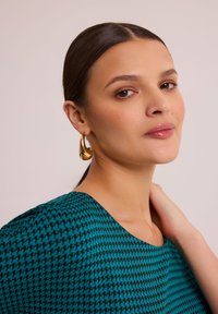 Tegołka w kolorze niebiesko-zielonym z wzorem w pepitkę, okrągłym dekoltem i krótkimi rękawami. Modelka nosi złote kolczyki w kształcie kropli. Gładka struktura materiału.