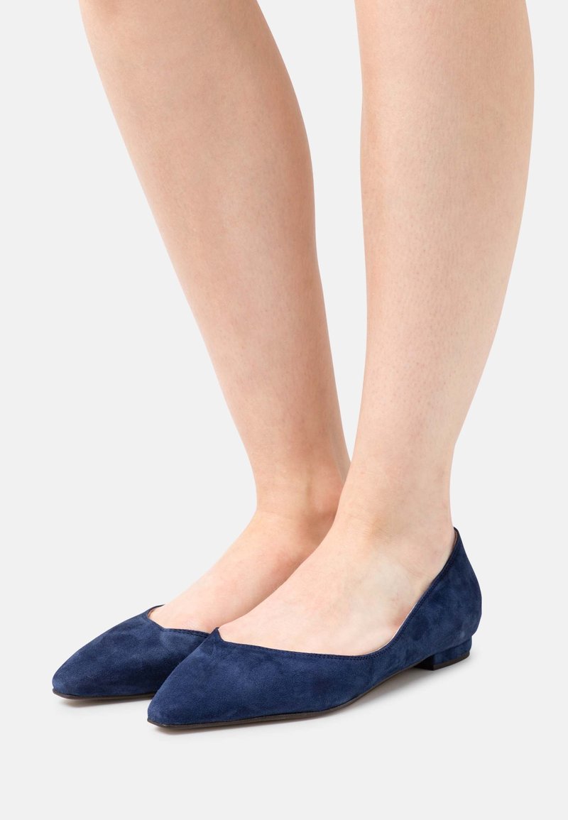 Paire de jambes portant des chaussures en daim bleu marine à bout pointu et à talon bas sur un fond clair.