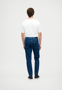 Jeans de denim azul escuro com um corte slim, apresentando um rótulo preto na cintura e dois bolsos traseiros. Combinados com uma t-shirt branca simples.