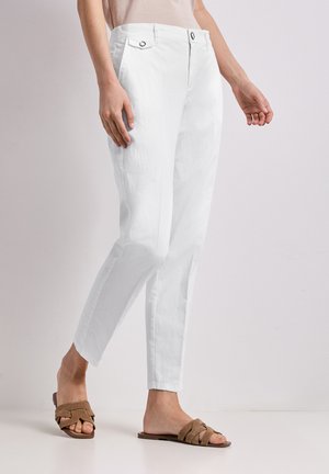 Femme portant un pantalon blanc slim associé à des sandales plates tressées marron, debout devant un fond clair uni.