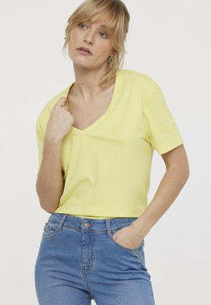 T-shirt jaune col V en coton doux, à manches courtes, avec un petit logo sur la poitrine gauche, associé à un jean bleu clair.