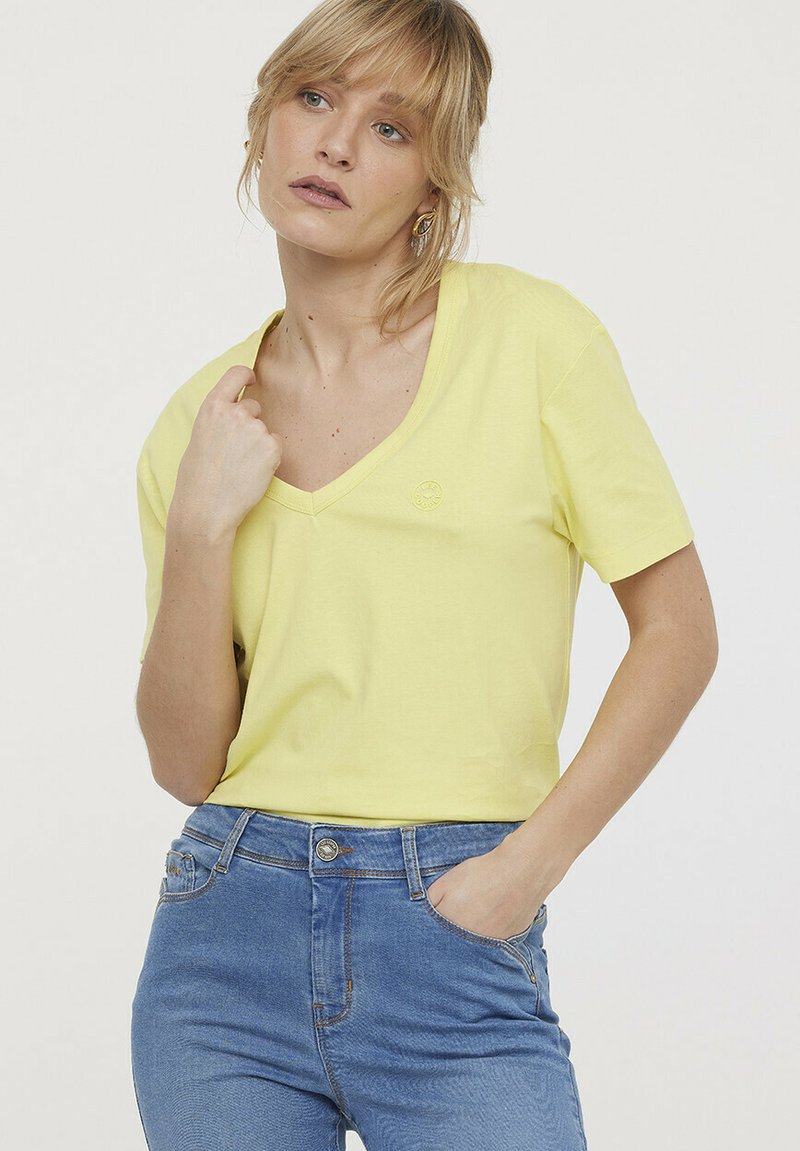 T-shirt jaune col V en coton doux, à manches courtes, avec un petit logo sur la poitrine gauche, associé à un jean bleu clair.