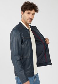 Marineblaue Lederjacke mit Reißverschluss, ausgestattet mit einer Fronttasche und rotem Akzentfutter, glatter Textur und lässigem Design.