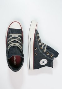 Baskets montantes en denim bleu avec des lacets beiges, semelles en caoutchouc et un logo circulaire sur le côté. Présente une bande rouge et blanche sur la semelle.