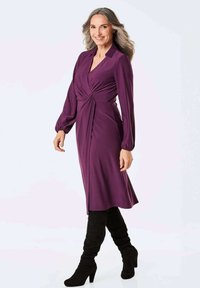 Langes lila Kleid mit einem gedrehten Vorderteil, knielangem Saum und weicher Textur. Getragen mit kniehohen schwarzen Stiefeln für einen Kontrast.