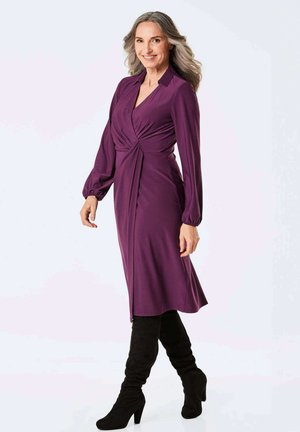 Langes lila Kleid mit einem gedrehten Vorderteil, knielangem Saum und weicher Textur. Getragen mit kniehohen schwarzen Stiefeln für einen Kontrast.