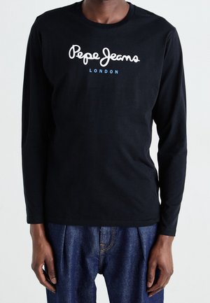 Personne portant une chemise noire à manches longues avec le texte "Pepe Jeans London" et un jean en denim bleu, debout sur un fond uni.