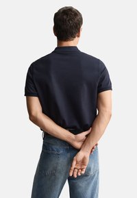 Granatowa koszulka polo z krótkimi rękawami, o fakturowanym materiale i klasycznym kołnierzykiem, zestawiona z wyblakłymi niebieskimi jeansami. Styl casual widziany od tyłu.