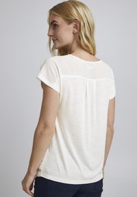 Cremefarbiges T-Shirt mit kurzen Ärmeln aus leichtem Stoff, versehen mit einem transparenten Overlay an der Rückseite und einem runden Halsausschnitt. Besitzt einen lockeren Schnitt.