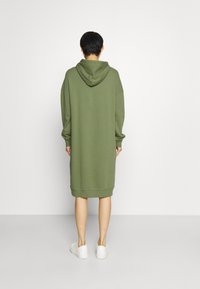 Abito verde oliva con cappuccio, maniche lunghe, vestibilità ampia e orlo a coste, realizzato in tessuto morbido; abbinato a sneakers bianche.