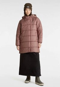 Gepolsterter, mauvefarbener Wintermantel mit Kapuze, elastischen Bündchen und Reißverschluss vorne. Kombiniert mit einem schwarzen langen Rock und grünen Stiefeln.