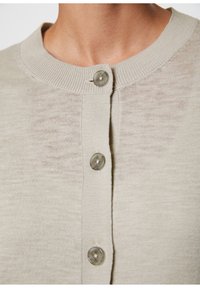 Beżowy kardigan z prążkowanym dekoltem i zapięciem na przodzie na przejrzyste guziki, o miękkiej i lekkiej fakturze.