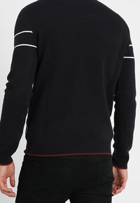 Pull en maille noire avec manches longues, accents rayés blancs sur les épaules et détail d'ourlet rouge.