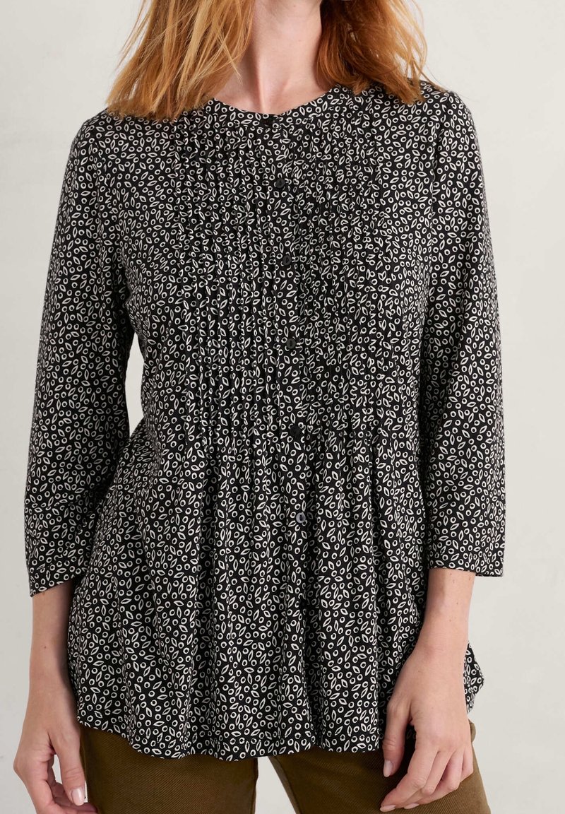 Femme portant un chemisier noir ample avec de petits motifs ovales blancs et des manches trois-quarts, assorti à un pantalon marron.