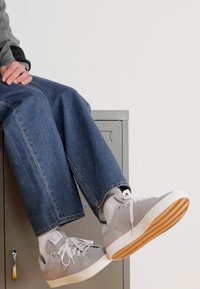 Sneaker grigi con suola bianca, realizzati in materiale liscio e con lacci bianchi. Indossati con jeans in denim blu larghi e calzini bianchi.